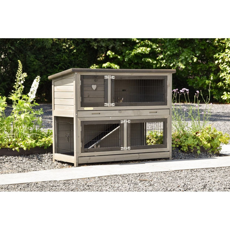 Zweistöckiger Holz-Kleintierstall mit Drahtgitter im Garten; obere Tür mit Herz-Dekor, darunter eine grössere Käfigebene, umgeben von Kies und Pflanzen.