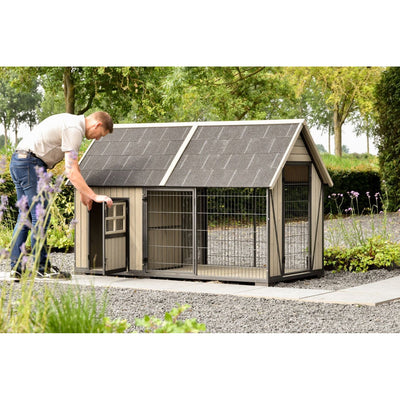 Mann öffnet die Tür einer grossen Hundehütte mit Auslauf im Garten; Holzrahmen, Drahtgitter, schräges Schindeldach, Kiesboden und grüne Bäume im Hintergrund.
