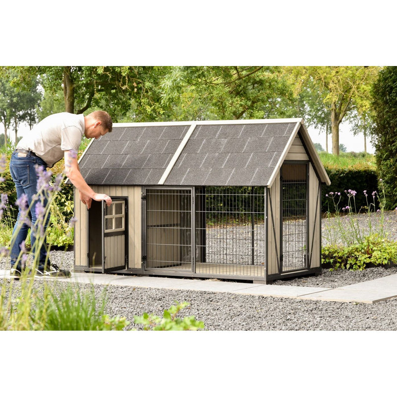 Mann öffnet die Tür einer grossen Hundehütte mit Auslauf im Garten; Holzrahmen, Drahtgitter, schräges Schindeldach, Kiesboden und grüne Bäume im Hintergrund.