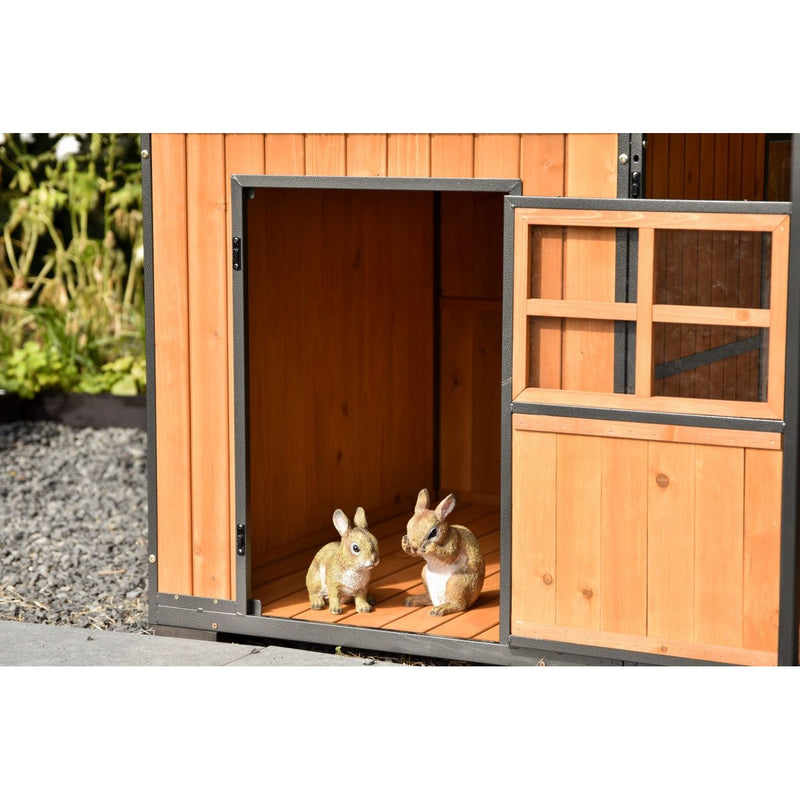 Zwei kleine Kaninchen-Figuren sitzen im offenen Gartenhaus aus orangefarbenem Holz mit schwarzem Metallrahmen; neben dem Eingang ist ein Fenster mit Holzrahmen sichtbar.