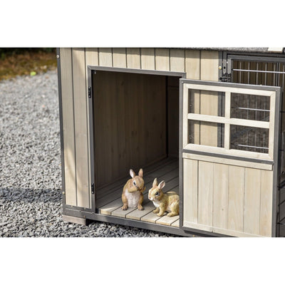 Zwei Hasen-Figuren stehen in einer offenen Holzhütte auf Kiesboden; die Hütte hat einen Metallrahmen und eine Tür mit Drahtgitter.