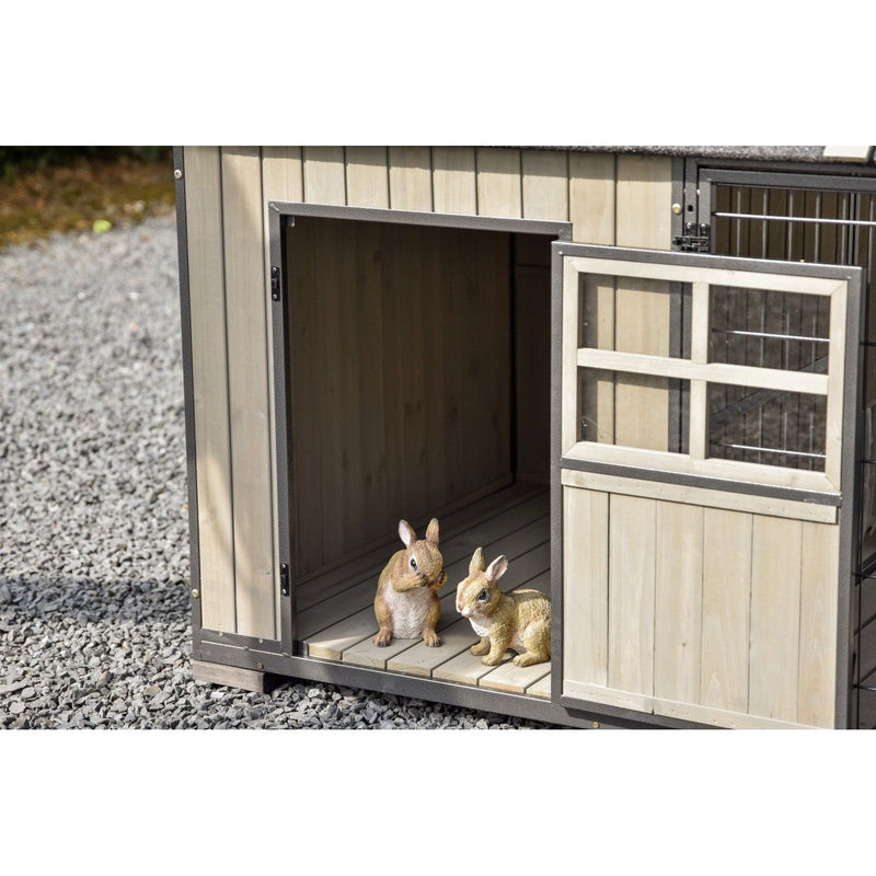 Zwei Hasen-Figuren stehen in einer offenen Holzhütte auf Kiesboden; die Hütte hat einen Metallrahmen und eine Tür mit Drahtgitter.