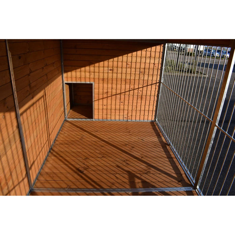 Innenraum eines holzverkleideten Geheges mit Metallgitter an der rechten Seite, kleinem quadratischen Eingang in der Rückwand, Holzboden und Schatten der Gitterstäbe.