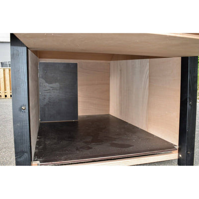 Offener Holzkasten-Innenraum aus Sperrholz mit schwarzer Bodenplatte und hellen Seitenwänden; Outdoor-Umgebung im Hintergrund.