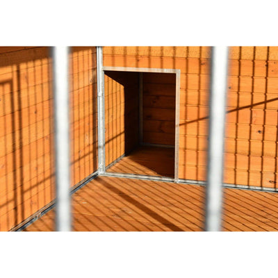 Orangefarbene Holzwand mit Metallrahmen, offener quadratischer Durchgang zu einem dunklen Innenraum; im Vordergrund Gitterstäbe werfen Schatten auf Boden und Wand.