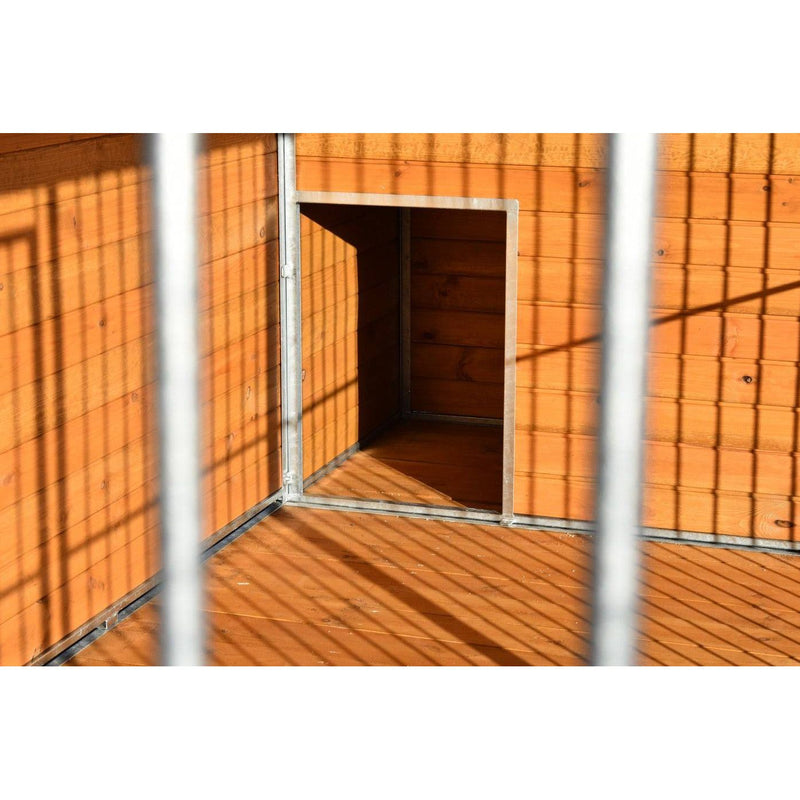 Orangefarbene Holzwand mit Metallrahmen, offener quadratischer Durchgang zu einem dunklen Innenraum; im Vordergrund Gitterstäbe werfen Schatten auf Boden und Wand.