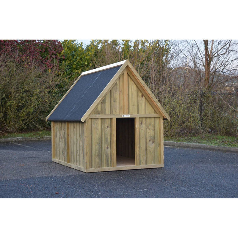 Kleine Holzhütte mit Satteldach aus Holzplanken auf einer asphaltierten Fläche, offenem Eingang, umgeben von Büschen und Bäumen.