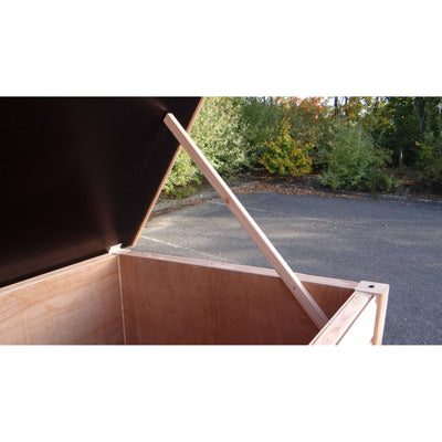 Geöffnete Holzkiste/Truhe mit Deckel, von einer diagonalen Holzstrebe gestützt, steht auf Asphalt neben einer Grünanlage.