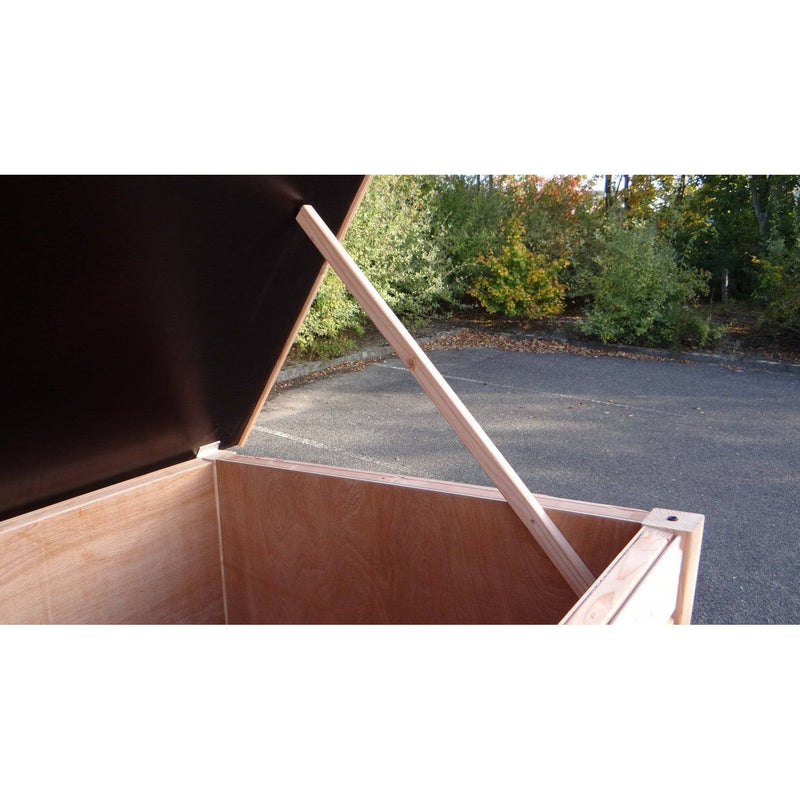 Geöffnete Holzkiste/Truhe mit Deckel, von einer diagonalen Holzstrebe gestützt, steht auf Asphalt neben einer Grünanlage.