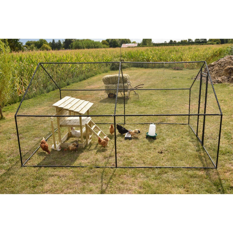 Grosses Hühnergehege aus Metallnetz mit Holzhäuschen und Leiter auf grasbewachsener Wiese; mehrere Hühner erkunden das Gelände, daneben Tränke und Futterautomat, Hintergrund Maisfeld und Heuhaufen.