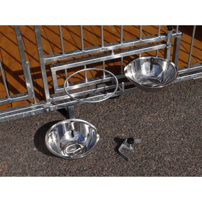 Zwei runde Edelstahl-Futternäpfe hängen an einem Metallzaun; ein weiterer Napf liegt auf dem Asphalt, daneben ein kleines Metallteil; Holzdecke im Hintergrund.